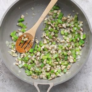 A pan with diced green bell pepper, onion, and mushroom being sautéed with a wooden spoon.