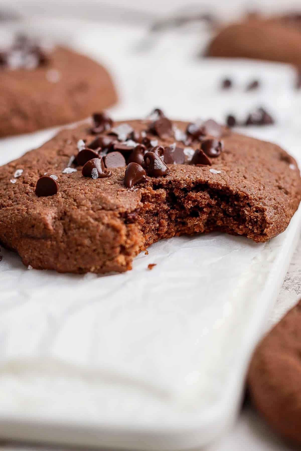 Close-up of a chocolate cookie with a bite taken out, topped with chocolate chips, resting on parchment paper.