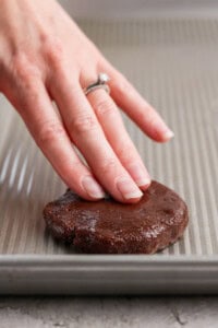 A hand presses down on a round chocolate cookie dough piece on a baking sheet.