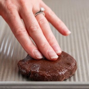 A hand presses down on a round chocolate cookie dough piece on a baking sheet.