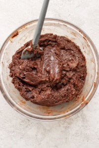 A glass bowl filled with thick, chocolate cookie dough and a gray spatula resting inside.