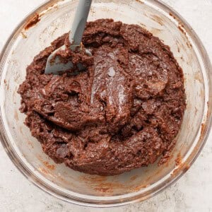 A glass bowl filled with thick, chocolate cookie dough and a gray spatula resting inside.