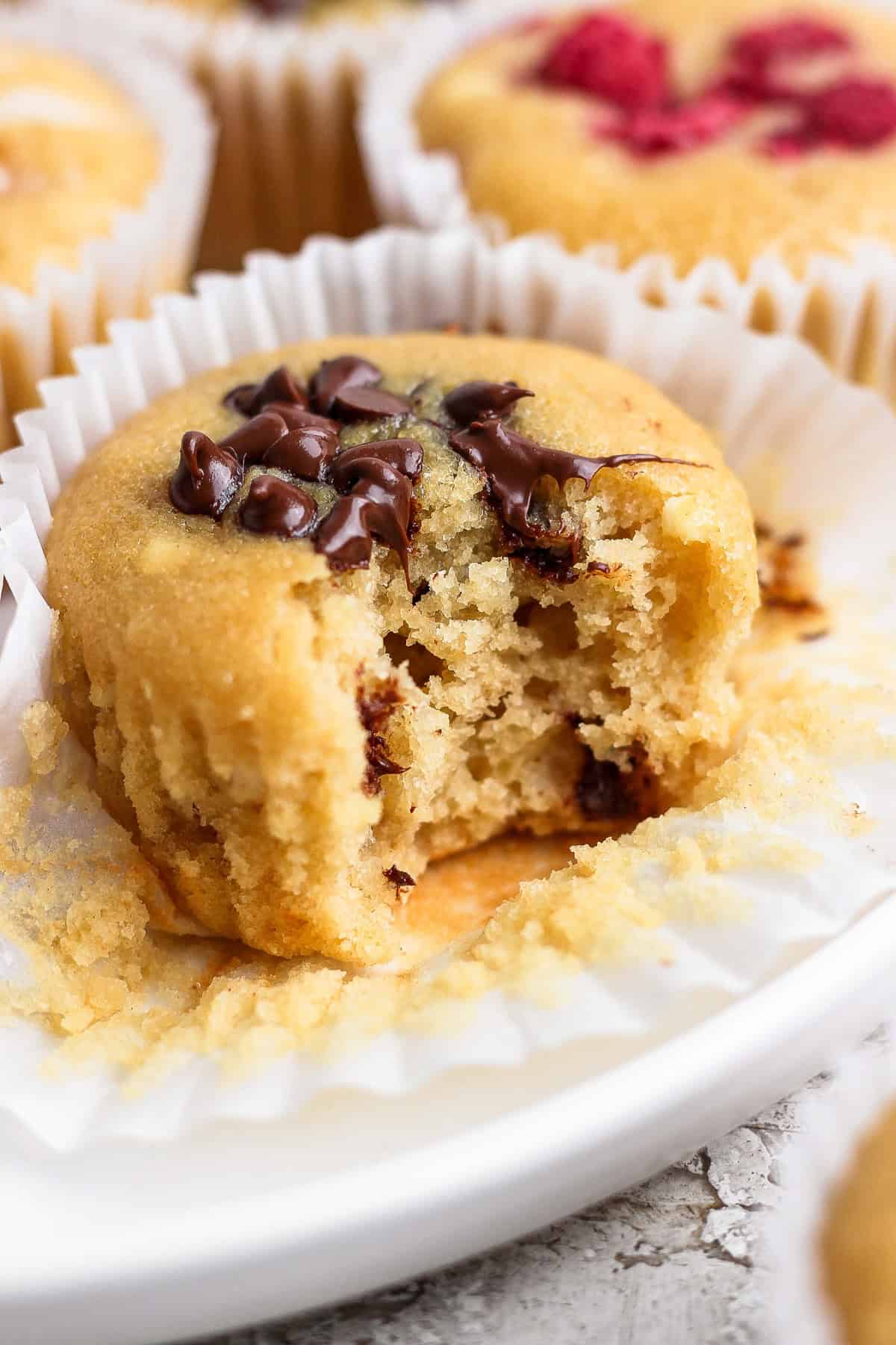 A muffin with chocolate chips on top, partially eaten, resting on a white paper liner.