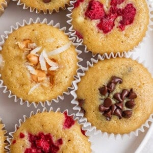 Assorted muffins topped with shredded coconut, chocolate chips, and raspberries in paper liners.