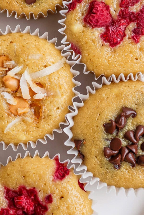 Assorted muffins topped with shredded coconut, chocolate chips, and raspberries in paper liners.