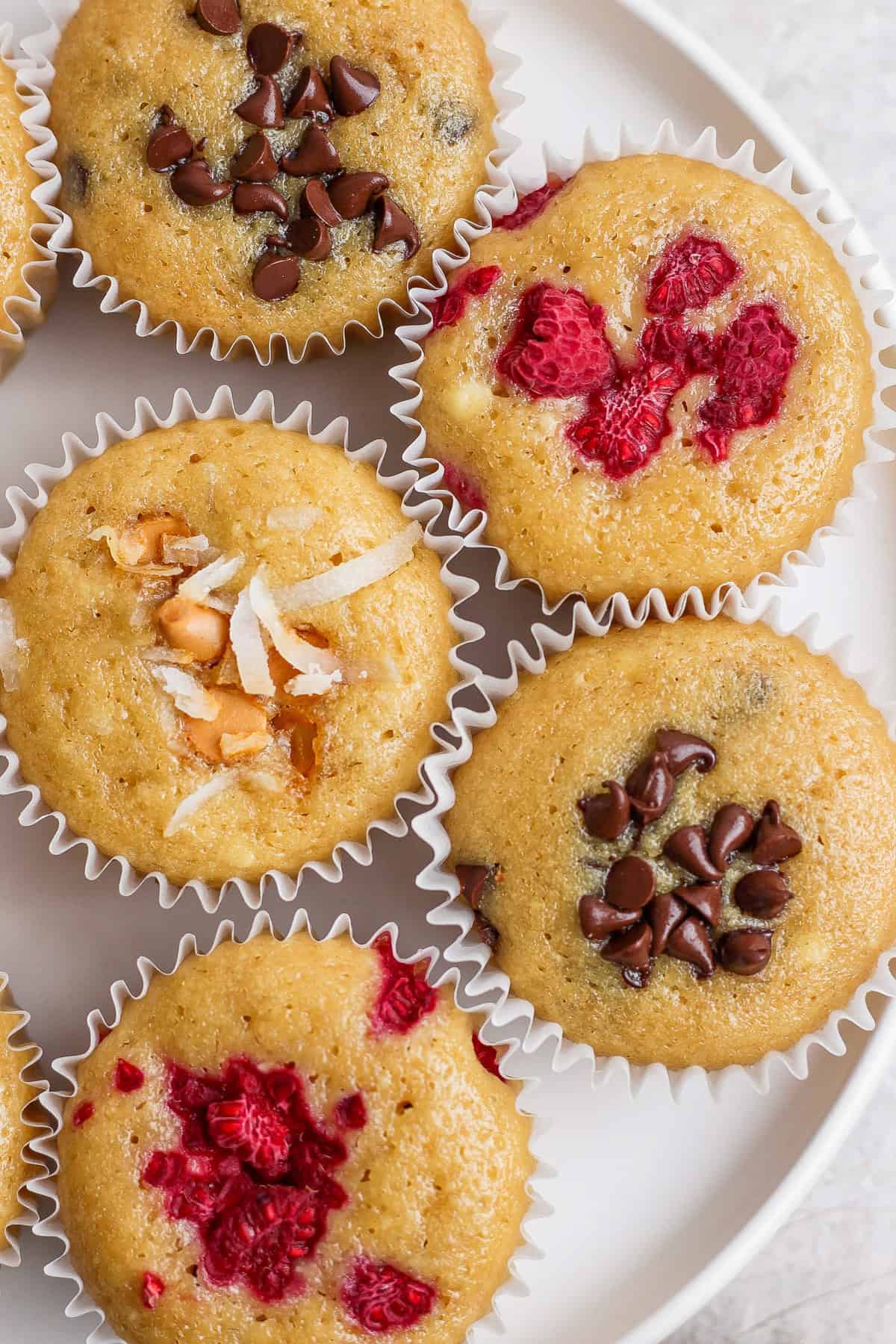 A plate with six muffins, some topped with chocolate chips, others with raspberries, and one with coconut and nuts, all in white paper liners.