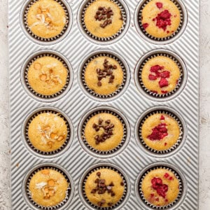 A muffin tray with 12 muffins, topped with almonds, chocolate chips, and raspberries.