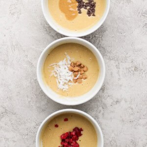 Three bowls of batter are topped with different ingredients: chocolate chips and peanut butter, shredded coconut and nuts, and red berries, arranged vertically on a textured surface.