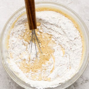 A glass bowl with a wooden-handled whisk mixing flour into a batter on a textured surface.