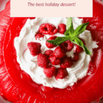 Red gelatin mold topped with whipped cream, raspberries, and mint leaves on a white plate.