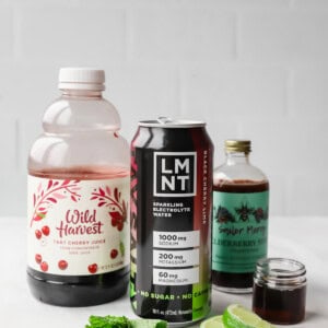 A bottle of tart cherry juice, a can of LMNT electrolyte drink, elderberry syrup, mint leaves, and lime slices on a white surface.