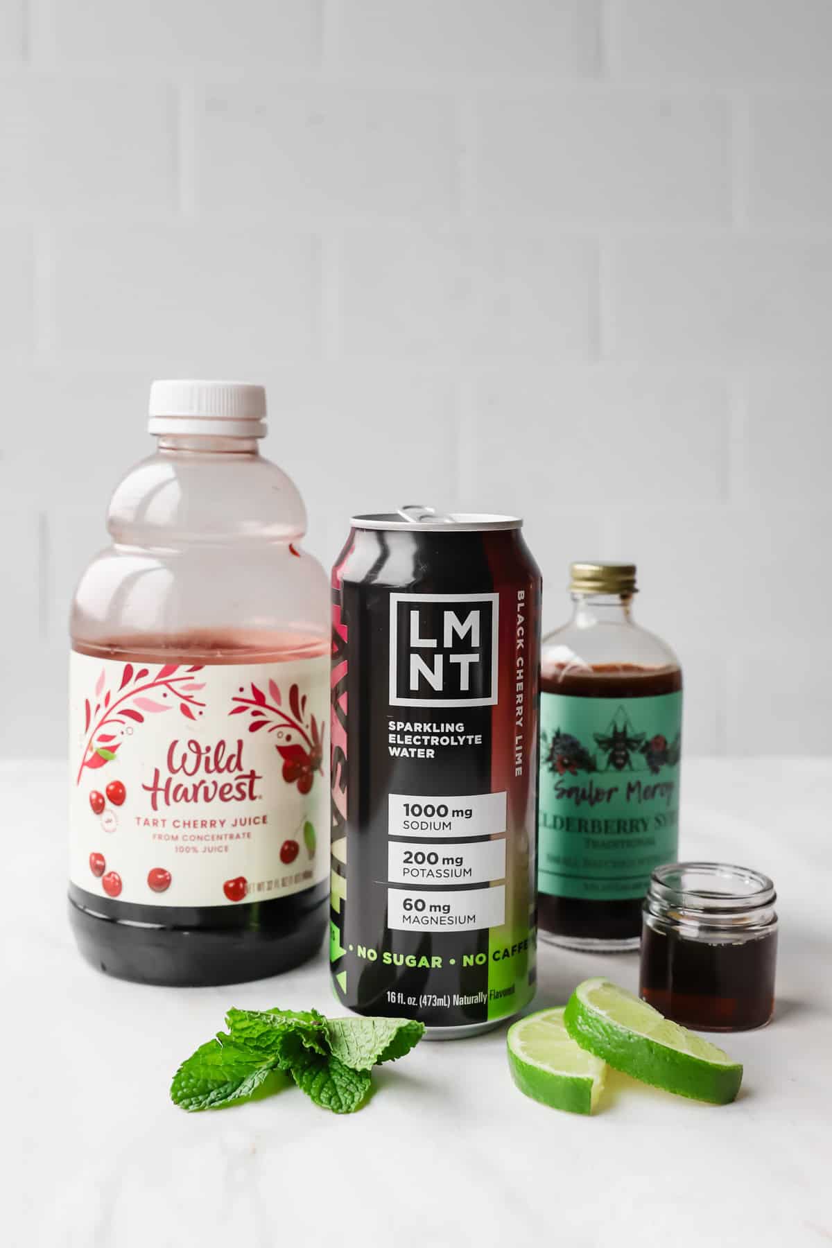 A bottle of tart cherry juice, a can of LMNT electrolyte drink, elderberry syrup, mint leaves, and lime slices on a white surface.