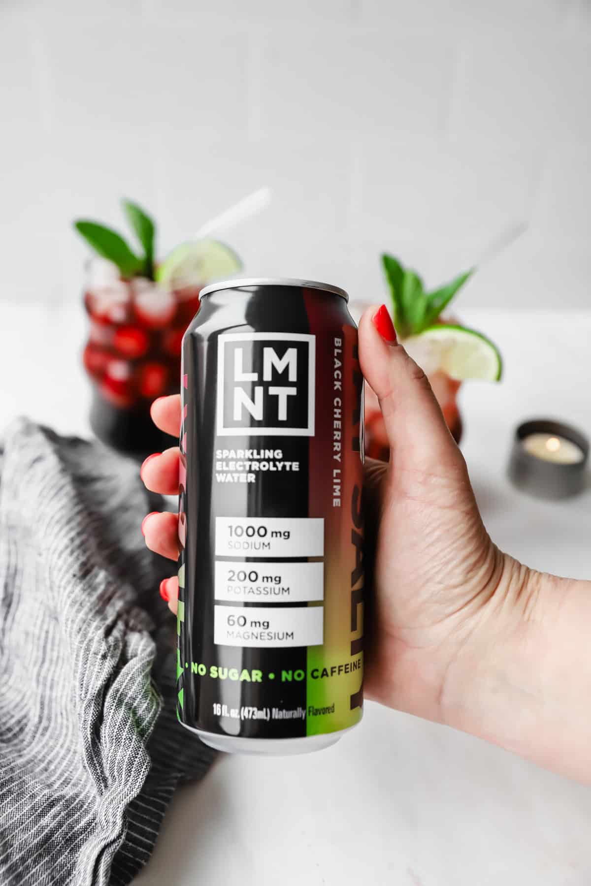 Hand holding a can of LMNT black cherry lime sparkling electrolyte water. Two glasses with red drinks and lime wedges are in the background.