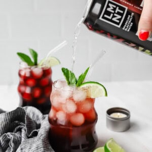 A hand pours a black cherry drink into a glass filled with ice, cherries, lime slices, and mint. Another similar drink is in the background, next to lime wedges and a small candle.