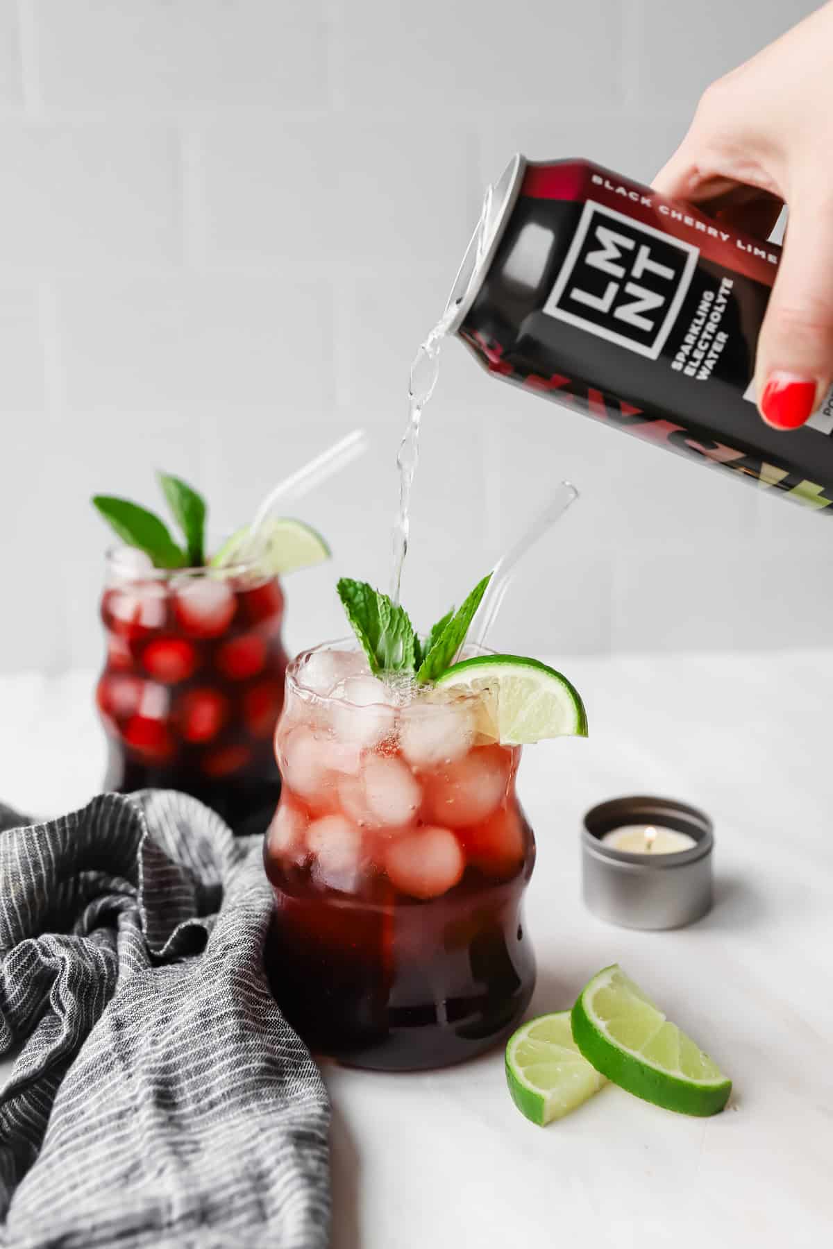 A hand pours a black cherry drink into a glass filled with ice, cherries, lime slices, and mint. Another similar drink is in the background, next to lime wedges and a small candle.