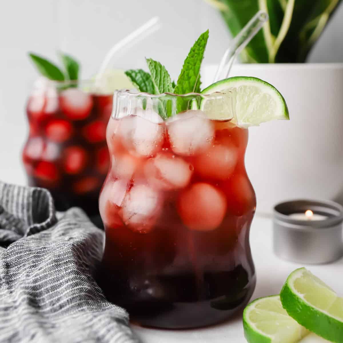 Two glasses of iced berry drink with lime slices and mint, placed on a table with a striped cloth. A small candle and lime wedges are nearby, with a potted plant in the background.