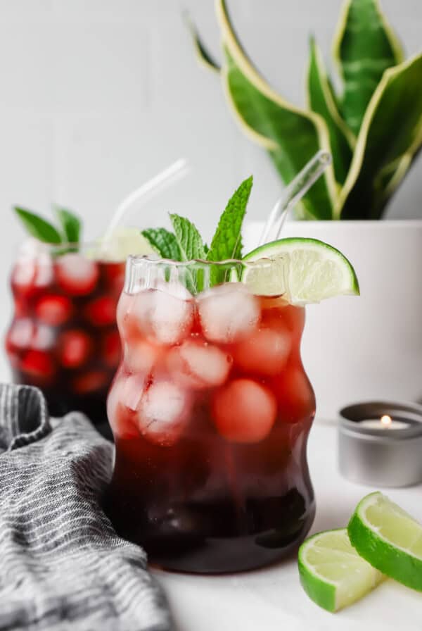 Two glasses of iced tea garnished with lime wedges and mint leaves. A potted plant and sliced limes are in the background.