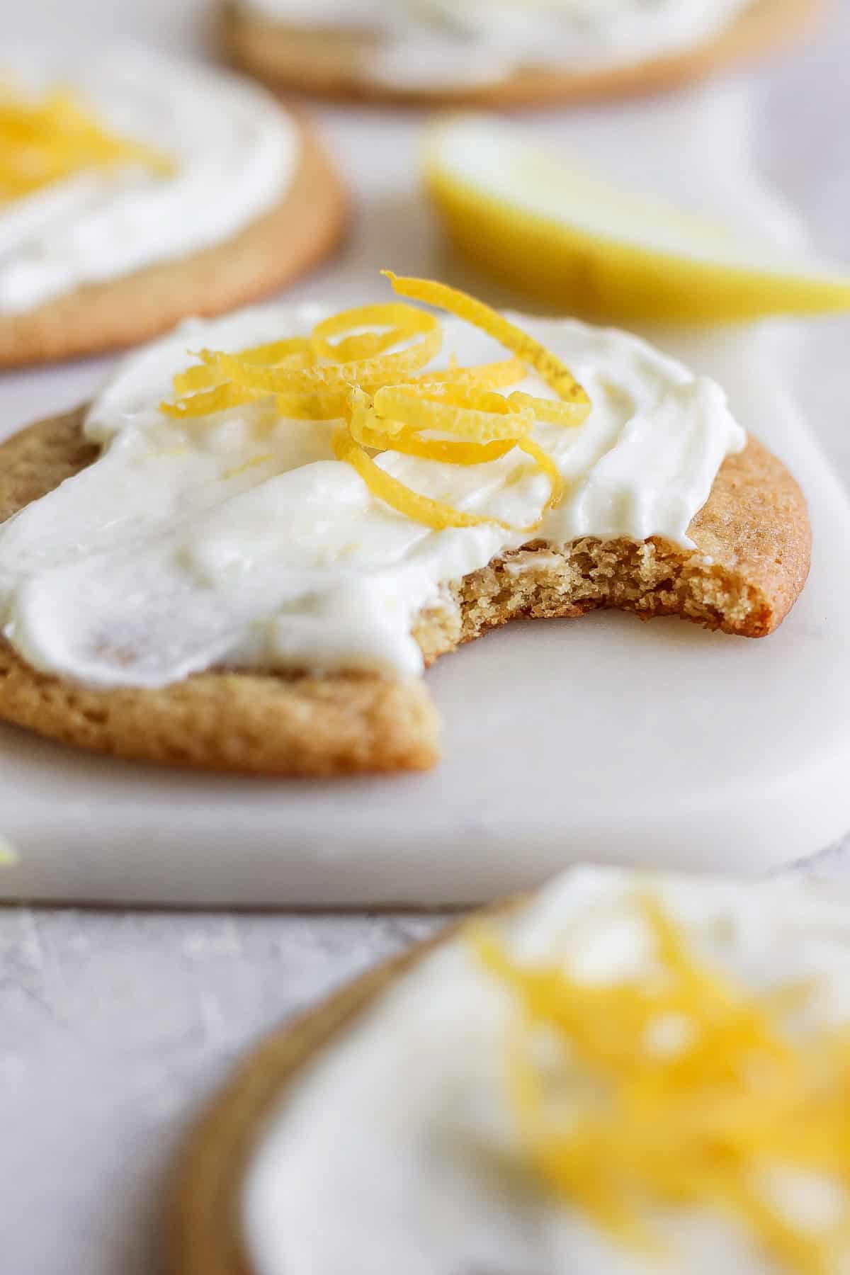 Close-up of a lemon cookie with cream cheese frosting and lemon zest on top. A bite is taken out of the cookie. A lemon wedge is visible in the background.