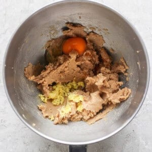 A mixing bowl brimming with cookie dough, a cracked egg, grated ginger, and hints of lemon zest prepares the stage for delightful lemon cookies with cream cheese frosting on a light textured surface.