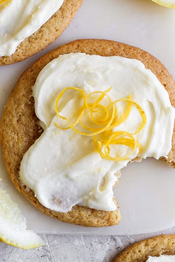 A lemon cookie with cream cheese frosting, topped with lemon zest, has a bite taken out of it. Slices of lemon are nearby on a light surface.