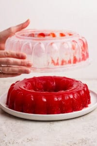 Hands removing a mold from a red gelatin dessert on a white plate.