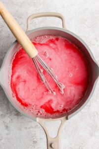 A whisk in a pot of frothy, red liquid on a light-colored surface.