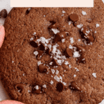 Close-up of a hand holding a chocolate peanut butter protein cookie topped with chocolate chips and sea salt. Text above reads "Chocolate PB Protein Cookies, 14g protein/cookie!.