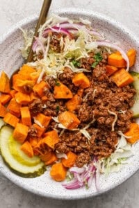 A bowl filled with ground beef, diced sweet potatoes, sliced pickles, shredded lettuce, and red onion slices, with a fork on the side.