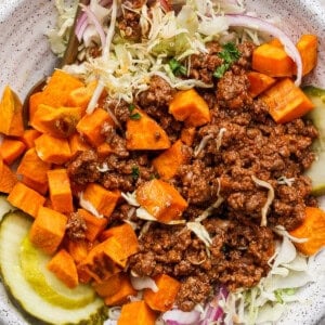 A bowl filled with ground beef, diced sweet potatoes, sliced pickles, shredded lettuce, and red onion slices, with a fork on the side.