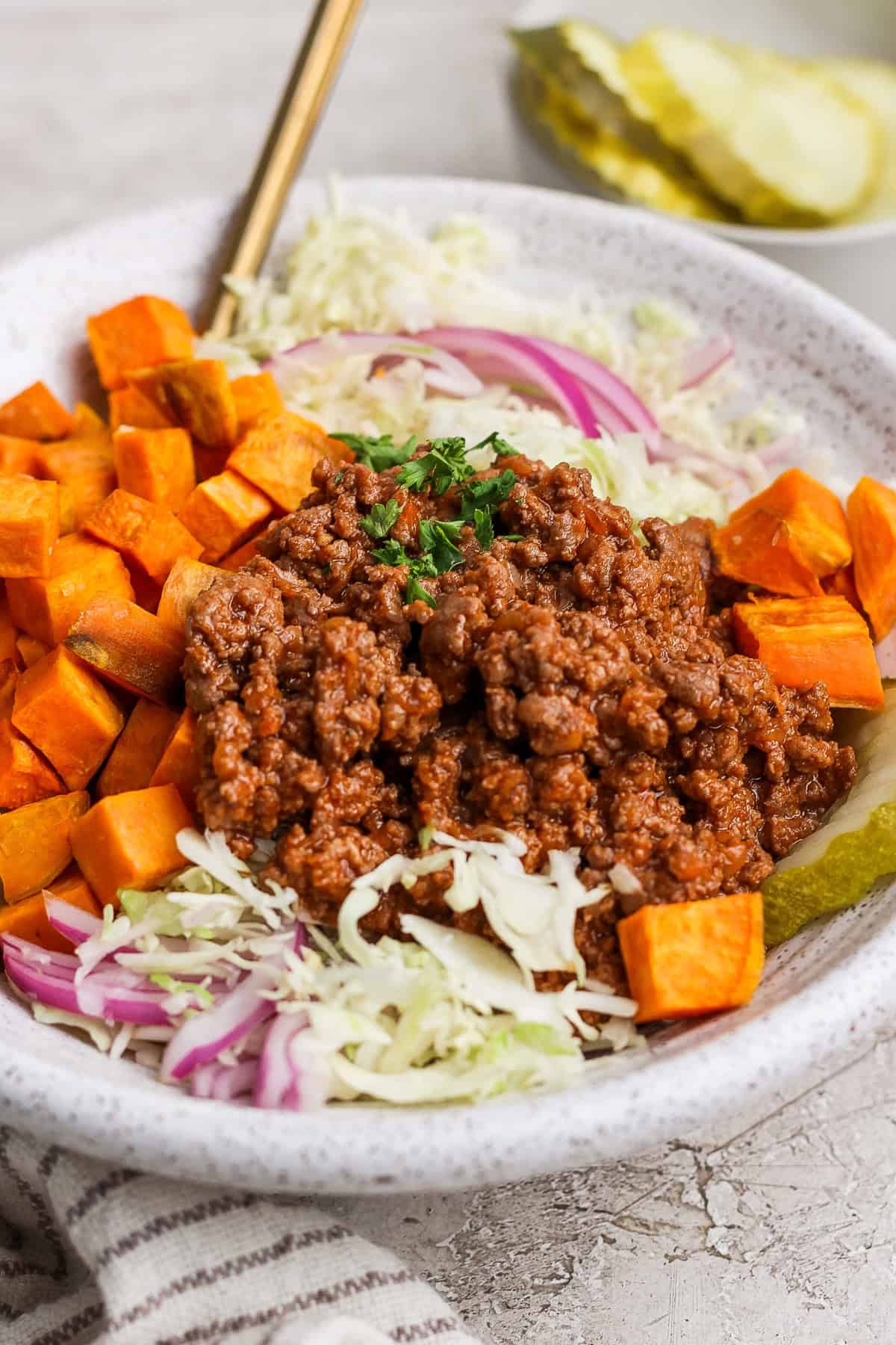 A bowl of salad with shredded cabbage, red onion, diced sweet potatoes, a scoop of seasoned ground meat, and garnished with parsley. Pickle slices are visible in the background.