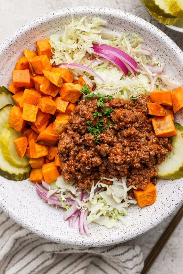 A bowl with ground meat, diced sweet potatoes, sliced pickles, shredded cabbage, and red onion.