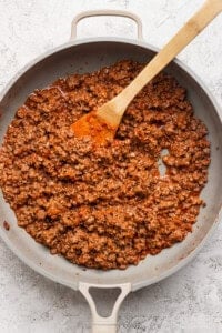 Cooked ground beef in a light-colored skillet with a wooden spatula, on a textured white surface.