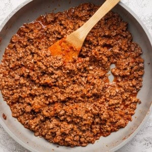 Cooked ground beef in a light-colored skillet with a wooden spatula, on a textured white surface.