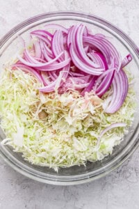 A glass bowl containing sliced red onions and shredded cabbage on a light gray surface.