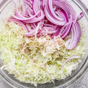 A glass bowl containing sliced red onions and shredded cabbage on a light gray surface.