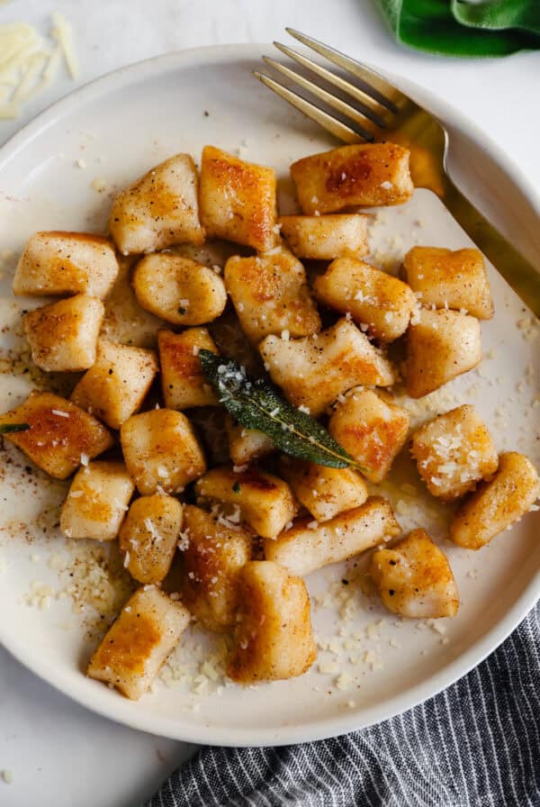 A plate of golden brown, cottage cheese gnocchi garnished with grated cheese and a sage leaf, accompanied by a fork.