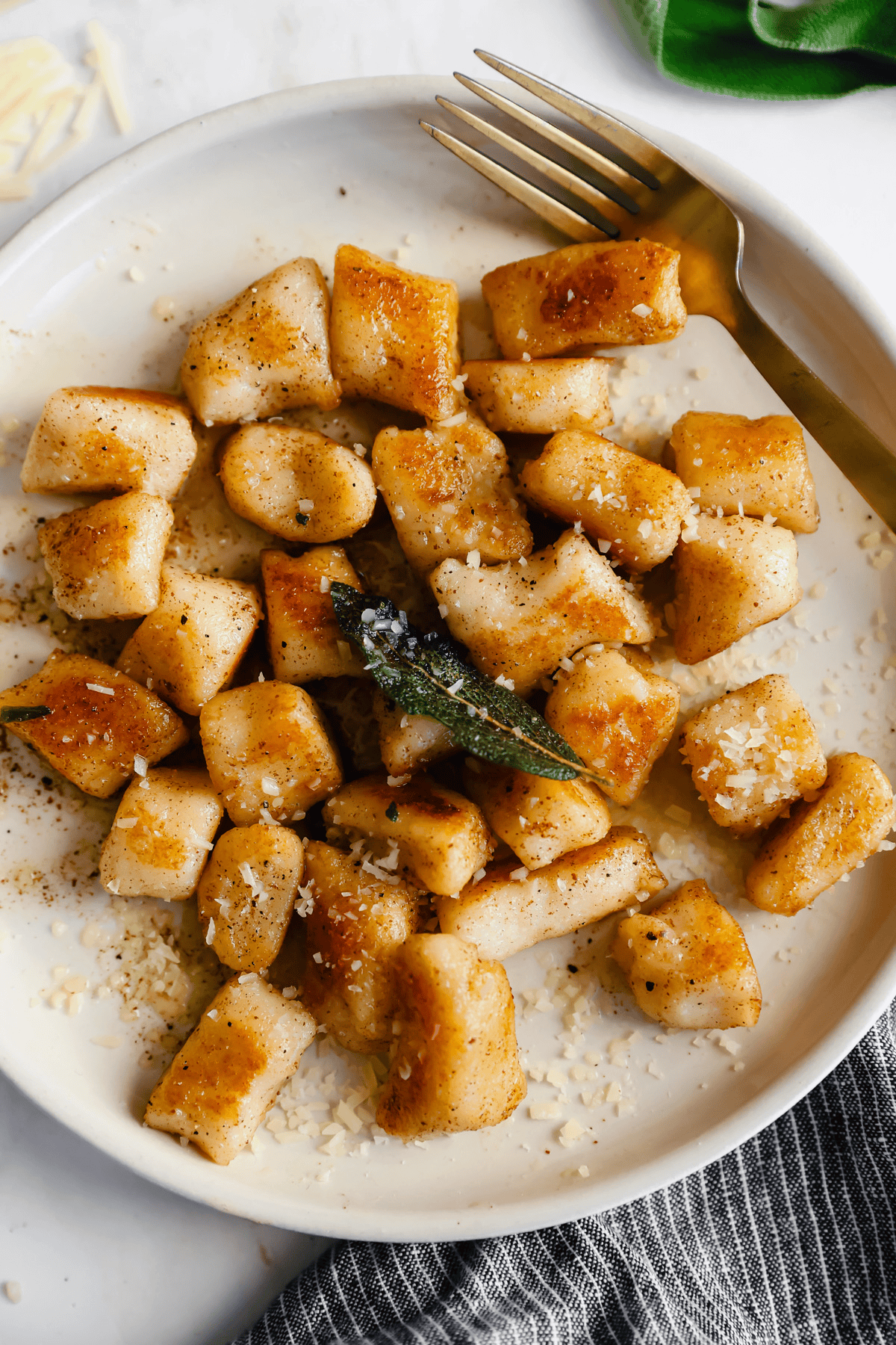 Plate of golden brown cottage cheese gnocchi with grated cheese and a sage leaf, accompanied by a fork, on a white background.