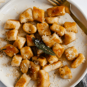 A plate of golden-brown cottage cheese gnocchi topped with fried sage and grated cheese, accompanied by a gold fork.