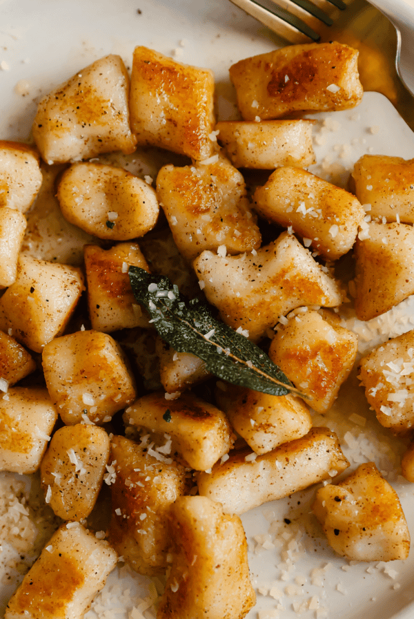 A plate of golden-brown cottage cheese gnocchi topped with fried sage and grated cheese, accompanied by a gold fork.