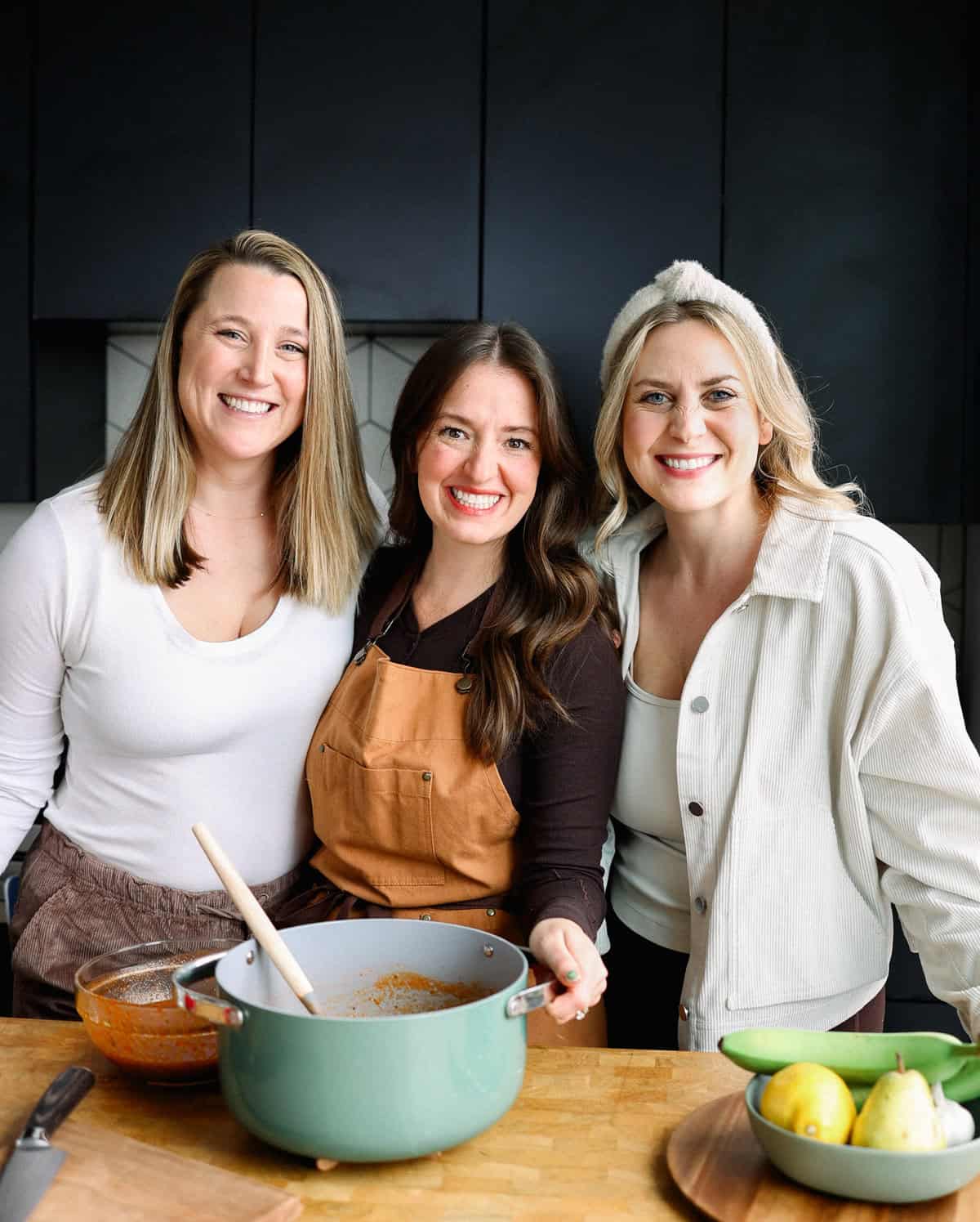 Three women smiling in a kitchen, embodying the spirit of fit foodie finds, stand behind a table adorned with a large pot, mixing bowl, and vibrant fruits.