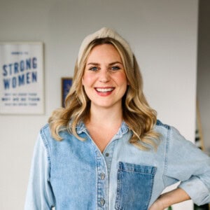 A smiling person with long blonde hair, wearing a denim shirt and a light hat, stands in front of a wall adorned with framed posters—it's the perfect spot for sharing about fit foodie finds.