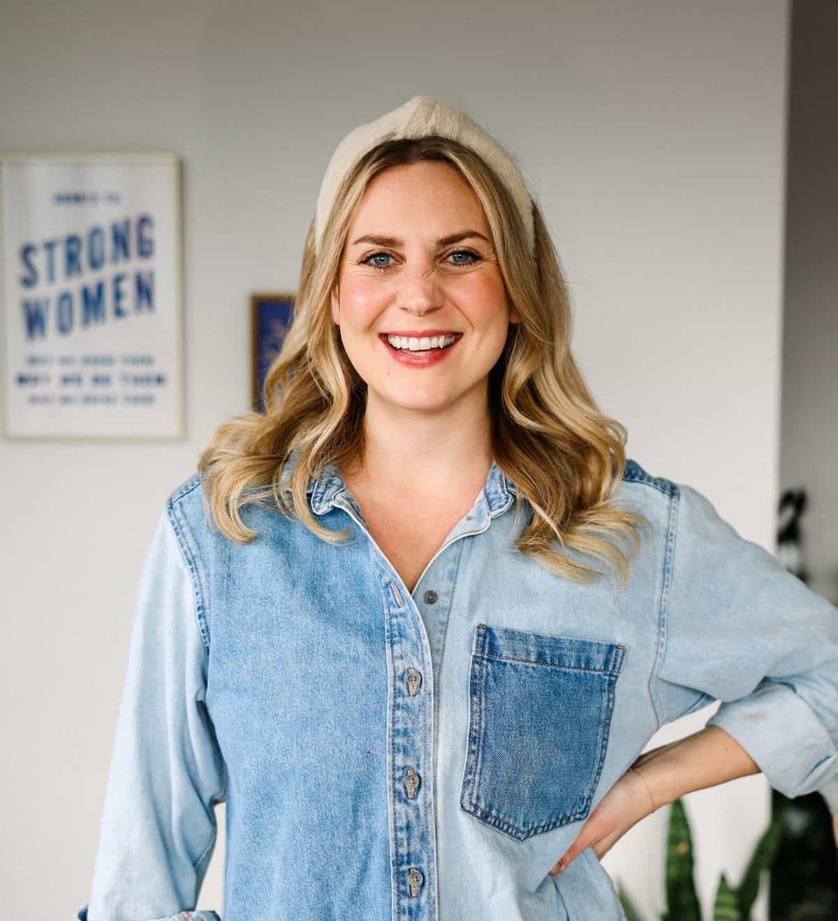 A woman with long blonde hair smiles, wearing a denim shirt and a light-colored headband, standing indoors, perhaps about to share her latest fit foodie finds.