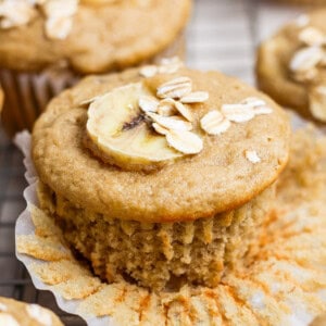 A banana muffin topped with a banana slice and oats, with a paper liner partially peeled back.