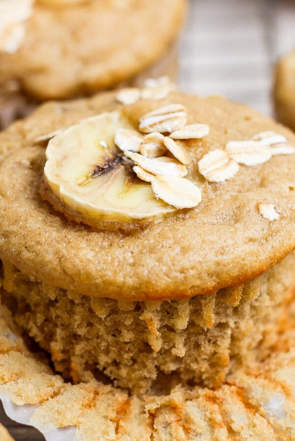 A banana muffin topped with a banana slice and oats, with a paper liner partially peeled back.