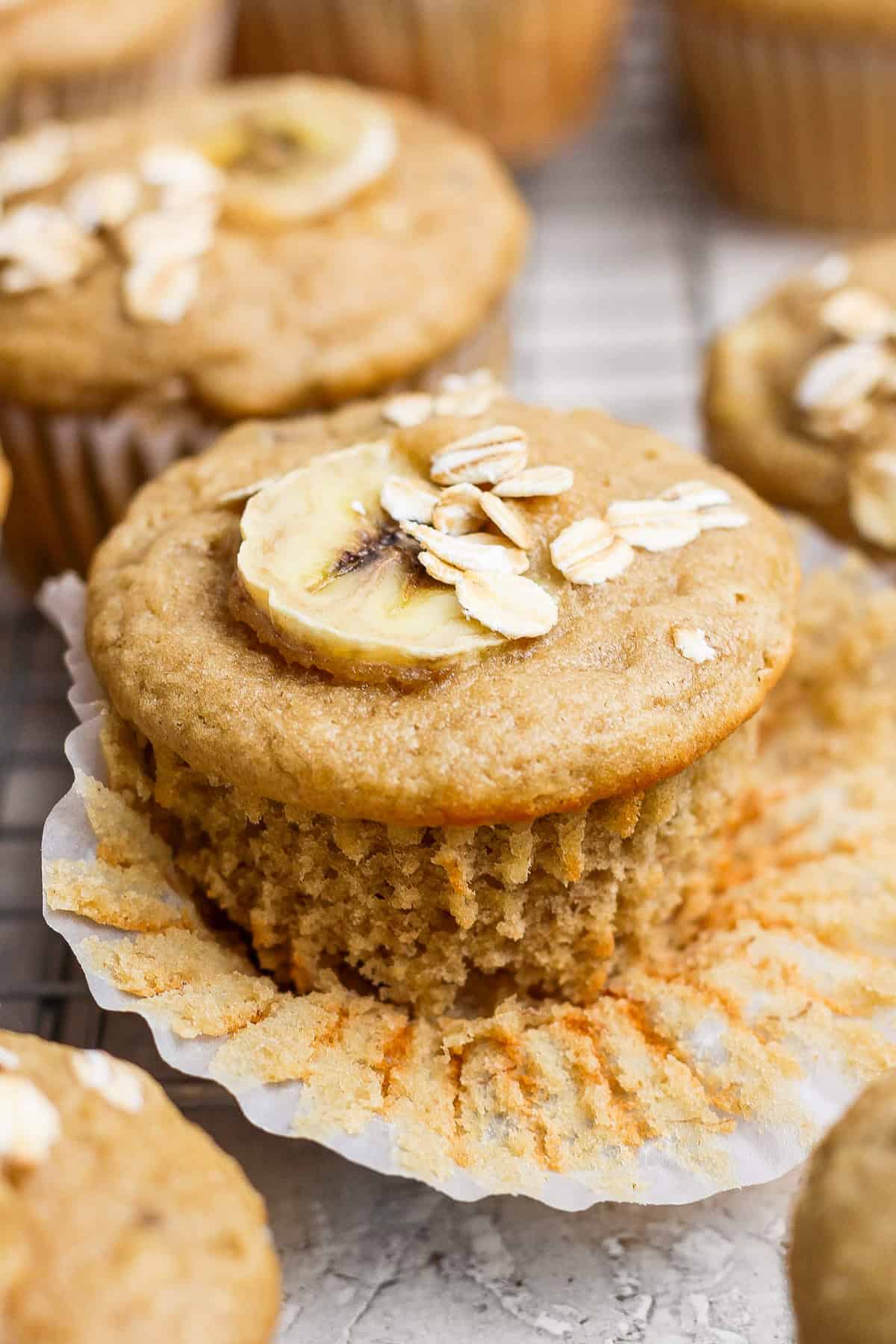 A banana muffin topped with a banana slice and oats, partially unwrapped on a wire rack.