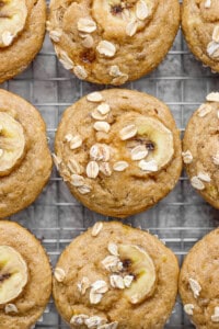 Banana oat muffins on a cooling rack, topped with banana slices and oats.