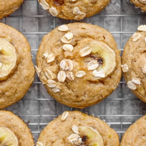 Banana oat muffins on a cooling rack, topped with banana slices and oats.