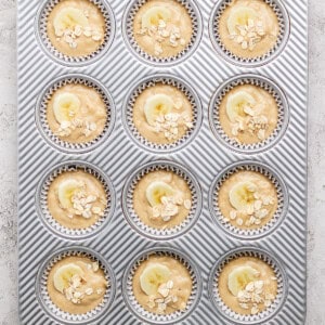 A muffin tray with twelve muffin batter portions, each topped with banana slices and oats, ready for baking.