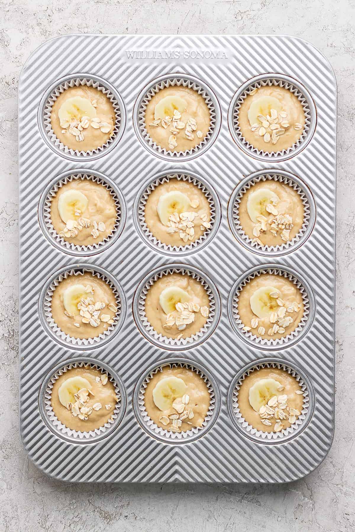 A muffin tray with twelve muffin batter portions, each topped with banana slices and oats, ready for baking.