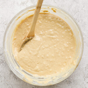 A glass bowl filled with creamy batter and a wooden spoon rests in it on a textured white surface.